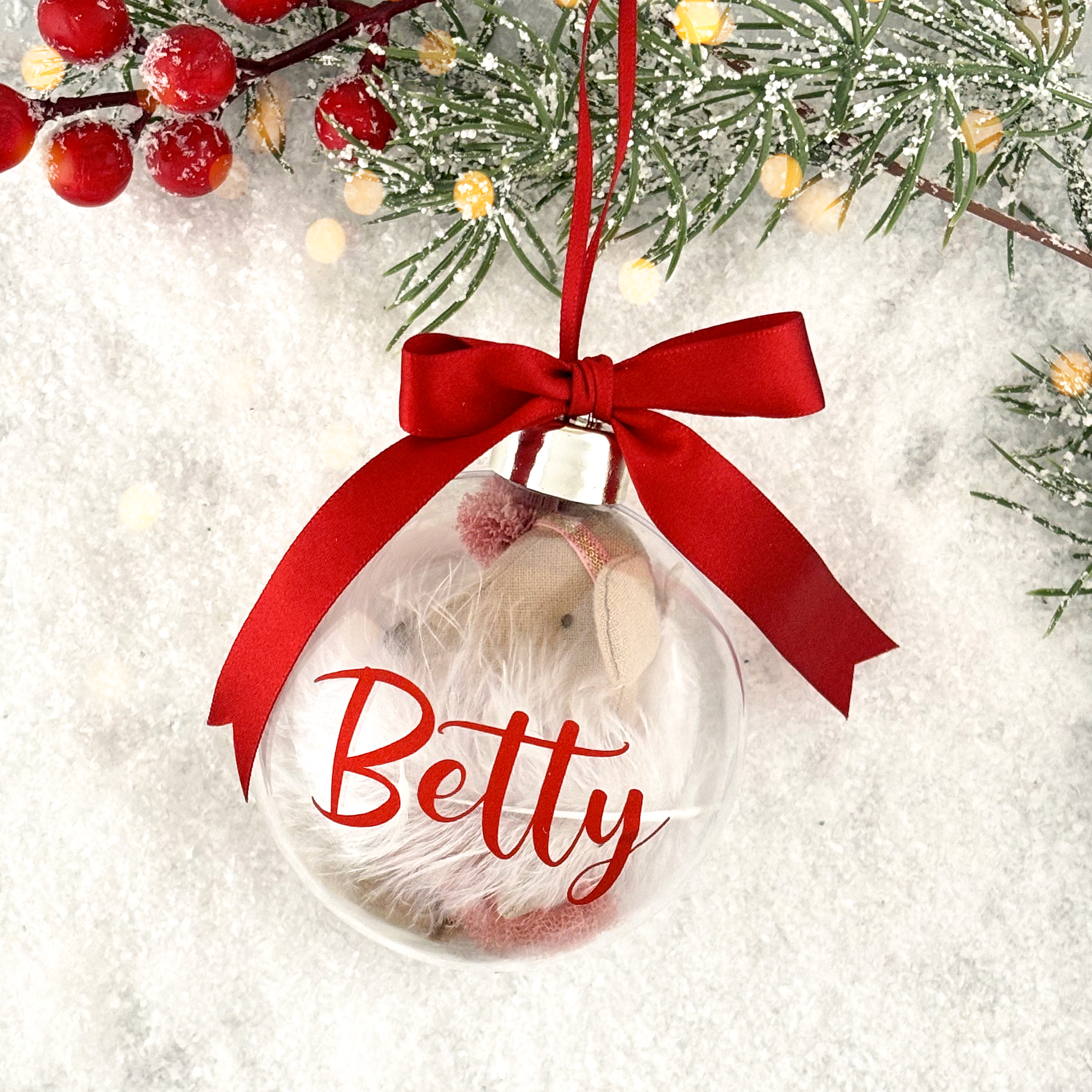 A personalized Christmas bauble with a red ribbon bow, featuring a white mouse nestled inside, surrounded by red and green artificial foliage and real holly berries on a snowy background.